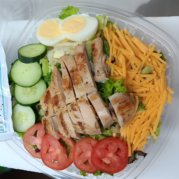 Grilled chicken salad with lettuce, tomatoes, cucumbers, hard-boiled egg, and shredded cheese in a plastic container.