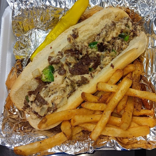 A Southern cheesesteak sandwich with fries and a pickle spear in a foil-lined container.