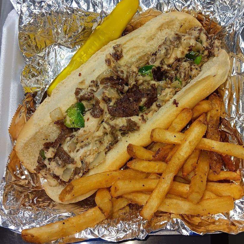 A Southern cheesesteak sandwich with fries and a pickle spear in a foil-lined container.