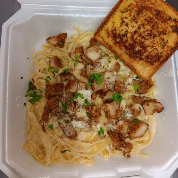 Creamy fettuccine pasta with seasoned chicken pieces and a slice of garlic bread in a takeout container.